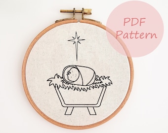 May include: A round embroidery hoop featuring a black line drawing of a baby in a manger, with a star above. The design is on a beige fabric, set in a wooden hoop. The text "PDF Pattern" is in a pink circle.