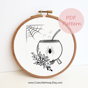 Op de afbeelding: Een zwart-witte borduurring met een Halloween-ontwerp. Het ontwerp omvat een spinnenweb, een ketel met een spin en bloemelementen. De tekst "PDF Pattern" staat in een roze cirkel. De website www.CuteLittleHoop.Etsy.com staat onderaan.