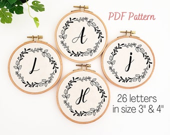May include: Four embroidery hoops with the letters A, J, L, and H, each surrounded by a black floral wreath. The text "PDF Pattern" and "26 letters in size 3" & 4"" are also visible.