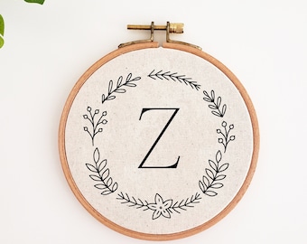 May include: A round embroidery hoop featuring the letter "Z" in black, centered on a beige fabric. The letter is surrounded by a wreath of black leaves and berries. The wooden hoop has a brass clasp.