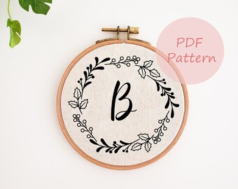 May include: A round embroidery hoop with a beige fabric featuring a black floral wreath design and the letter "B". The text "PDF Pattern" is in a pink circle. The hoop is made of wood.