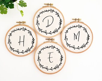 May include: Four embroidery hoops with the letters "D", "H", "M", and "E" stitched in black script. Each letter is surrounded by a black floral wreath on a beige fabric background. The hoops are made of wood and have brass hardware.