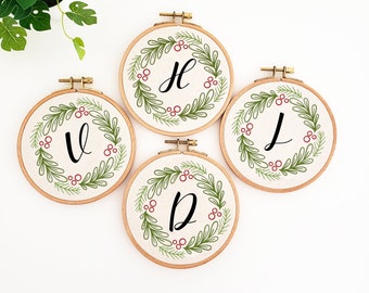 May include: Four embroidery hoops with the letters V, H, J, and D. Each hoop features a green leaf wreath design with red berries and a black letter. The hoops are set against a white background.