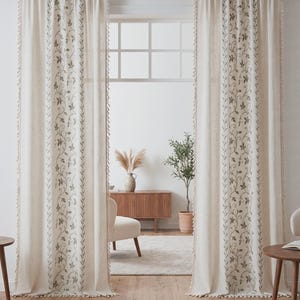 Peut inclure: Une paire de rideaux blanc cassé suspendus à une tringle sombre, présentant un panneau vertical avec un motif de vigne et de feuilles vertes. Les bords des rideaux sont ornés de petites franges claires. Le texte WarmOurLinen est en bas.