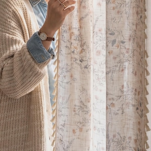 Peut inclure: Un panneau de rideau semi-transparent orné d'un imprimé botanique délicat avec des motifs floraux et végétaux aux tons doux de bleu et d'orange. Le tissu présente un tissage texturé et une bordure verticale de petites franges crème.