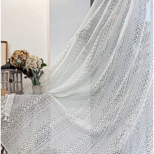 以下が含まれることがあります： 幾何学模様の白いクロシェ生地。生地はフリンジの縁取りがあり、花瓶の花が置かれたテーブルにかけられています。