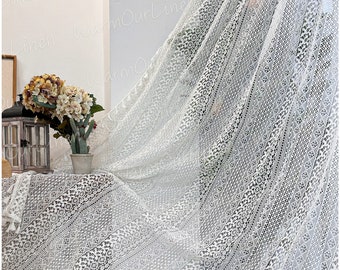 May include: White crochet fabric with a geometric pattern. The fabric has a fringe border and is draped over a table with a vase of flowers.