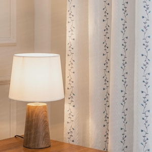 May include: A table lamp with a conical wood-grain base and a white fabric shade, positioned next to white curtains featuring vertical embroidered blue floral vines. The text WarmOurLinen is visible in the bottom right corner of the image.