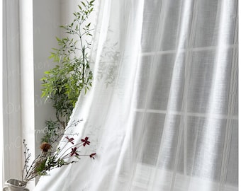 May include: Sheer white linen curtains with vertical stripes hang in front of a window. The curtains are partially drawn, allowing light to filter through. A vase with flowers and greenery sits in front of the curtains.