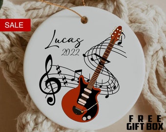 May include: White ceramic ornament with a red and black guitar design, musical notes, and the name "Lucas" with the year "2022". The ornament has a gold string for hanging.
