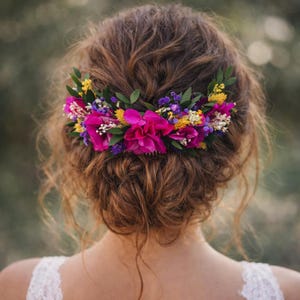 Könnte beinhalten: Ein Haarkamm mit einem lebendigen Arrangement aus magentafarbenen Blumen, kleinen violetten und gelben Blüten sowie grünen Blättern. Das Accessoire ist in welligem braunem Haar befestigt und bildet ein farbenfrohes florales Arrangement.