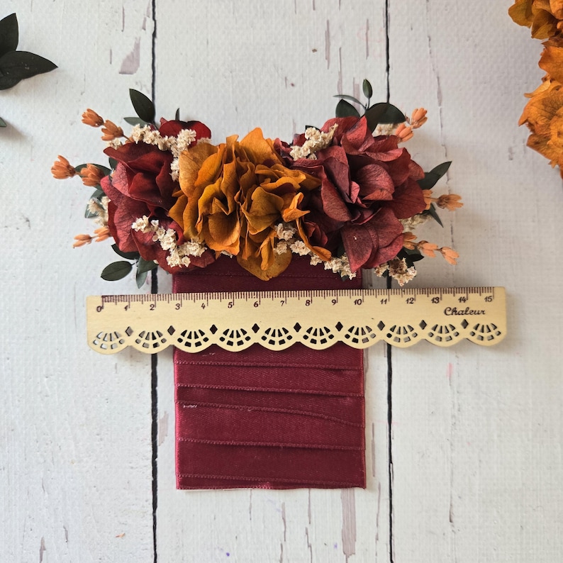 May include: A burgundy satin ribbon with a decorative lace trim and a floral arrangement of dried orange and burgundy flowers. The flowers are arranged in a cluster and are accented with small white flowers. A ruler is placed below the ribbon to show the size.