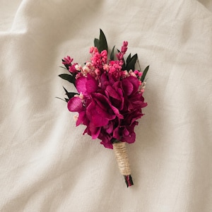May include: A boutonniere featuring deep magenta hydrangea petals, bright pink baby's breath, and dark green foliage. The stems are tightly wrapped in natural tan twine, creating a compact floral arrangement with a distinct color contrast.