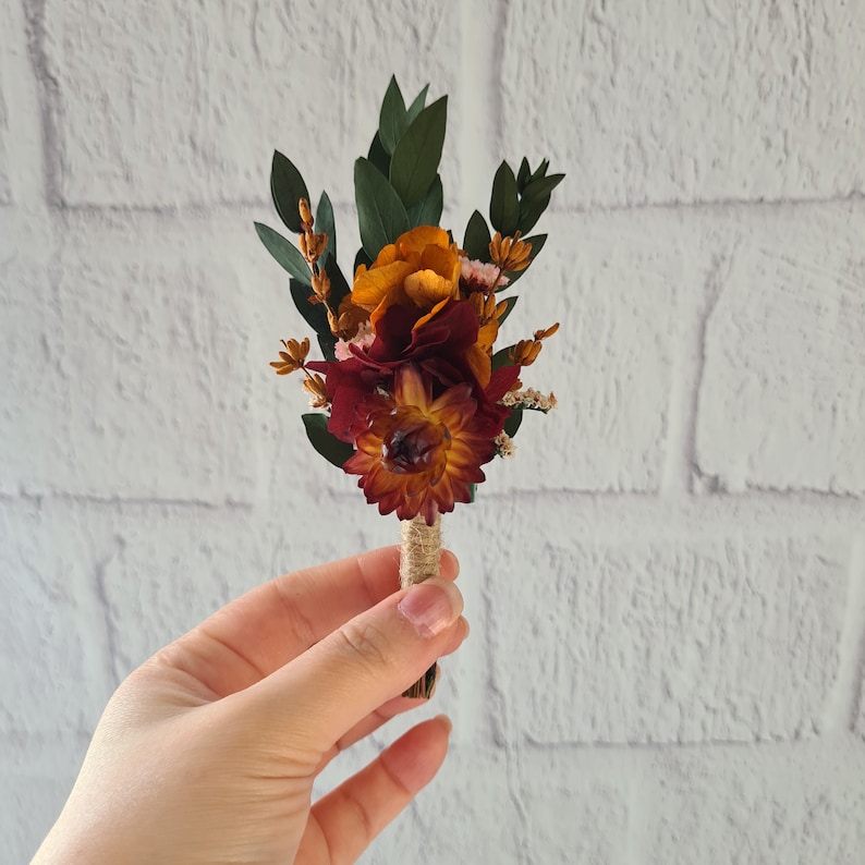May include: A small bouquet of dried flowers in shades of orange, red, and green, tied together with twine. The bouquet is held in a hand.