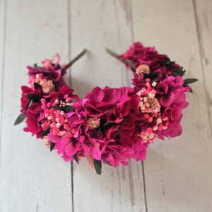 May include: A headband adorned with a dense arrangement of vibrant fuchsia and magenta hydrangea petals, interspersed with small clusters of light pink baby's breath and dark green leaves, creating a full, rounded floral crown design.