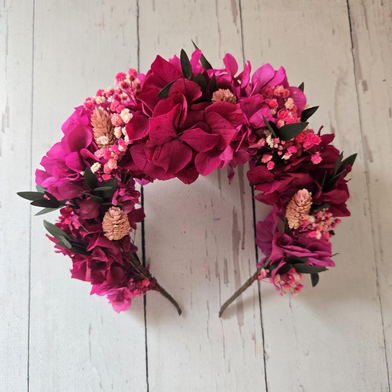 May include: A floral headband featuring vibrant magenta hydrangea petals, small clusters of light pink baby's breath, pale tan dried wheat-like stalks, and dark green leaves arranged in a dense, semi-circular crown design on a thin wire frame.