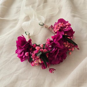 May include: A floral crown featuring deep magenta flowers and small pink baby's breath accents. The arrangement is secured on a base with thin, light-colored ribbon ties extending from the ends, creating a circular shape for head adornment.
