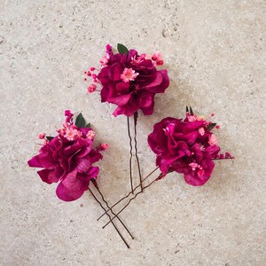 May include: Three hair pins featuring clusters of deep magenta flowers and small light pink blossoms. Each pin has a metallic U-shaped base topped with dark green leaves and vibrant, layered petals, creating a cohesive floral hair accessory set.