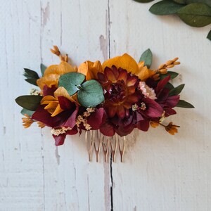 May include: A burgundy and gold floral hair comb with a gold metal comb. The comb is decorated with dried flowers, including burgundy hydrangeas, gold roses, and green eucalyptus leaves.