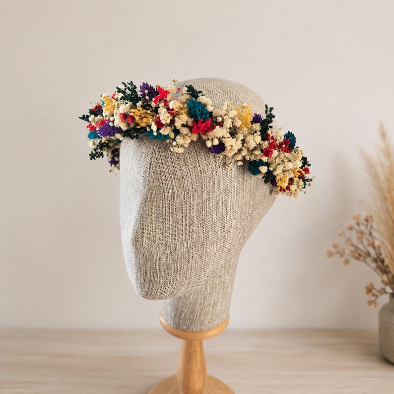 May include: A floral crown featuring a dense arrangement of dried flowers in vibrant shades of red, yellow, teal, and purple, interspersed with small white baby's breath blossoms. The circular headpiece is displayed on a neutral-toned mannequin head.