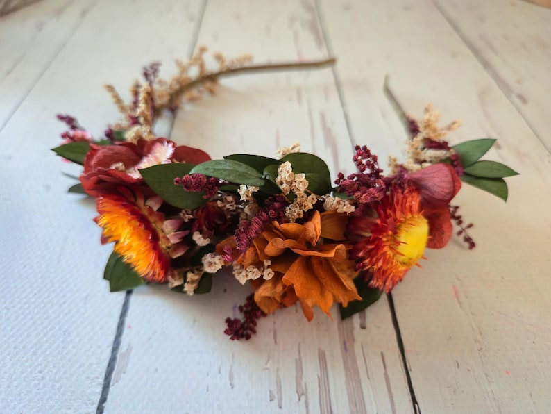 May include: A floral headband with dried flowers in shades of orange, red, and yellow. The headband is made with a wire base and is decorated with small sprigs of dried flowers.