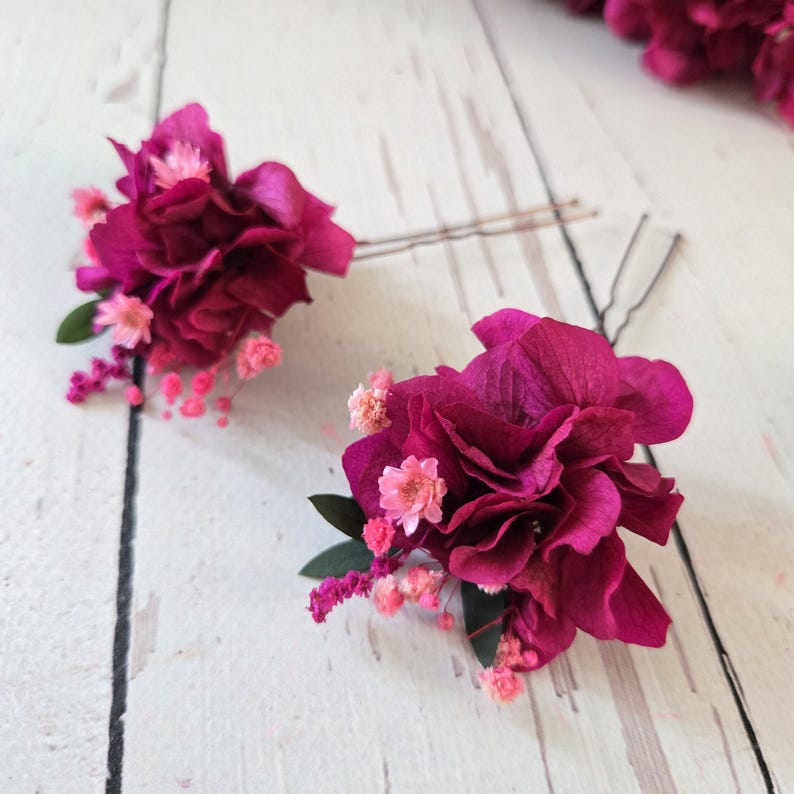 May include: Two hair pins featuring clusters of deep magenta hydrangea petals, small light pink filler flowers, and tiny green leaves. Each floral arrangement is securely attached to a thin, dark metal U-shaped hair pin for styling hair.