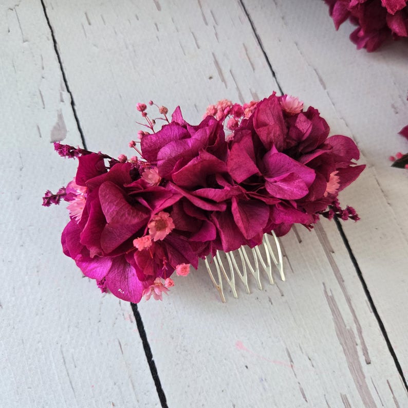 May include: A hair comb featuring a cluster of deep magenta hydrangea petals and small light pink baby's breath flowers. The decorative floral arrangement is attached to a silver-toned metal comb with multiple prongs for securing into hair.