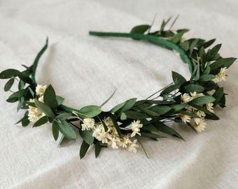 May include: A circular headband featuring a thin green frame adorned with small, pointed dark green leaves and clusters of tiny, off-white flowers distributed evenly along the entire length of the accessory, creating a natural, botanical appearance.