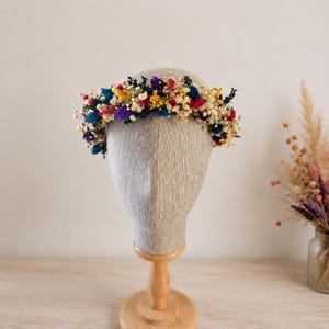 May include: A floral crown featuring a vibrant mix of dried flowers in shades of cream, yellow, red, purple, and blue. The circular arrangement is displayed on a neutral-toned mannequin head, showcasing a dense collection of small blossoms.