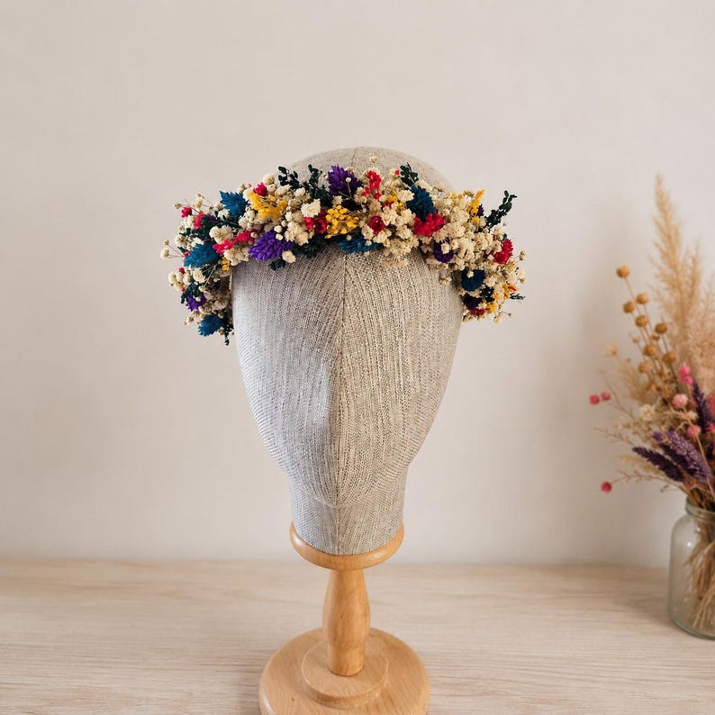 May include: A floral crown featuring a vibrant mix of dried flowers in shades of cream, yellow, red, purple, and blue. The circular arrangement is displayed on a neutral-toned mannequin head, showcasing a dense collection of small blossoms.