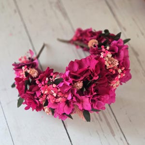 May include: A headband adorned with a dense arrangement of vibrant fuchsia and magenta hydrangea petals, interspersed with small clusters of light pink baby's breath and dark green leaves, creating a full, textured floral crown design.