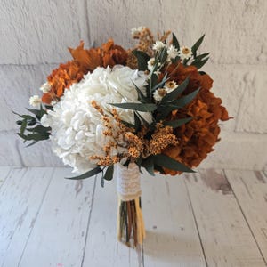 May include: A dried flower bouquet with white hydrangeas, orange flowers, and green eucalyptus leaves. The stems are bound with twine and lace. The arrangement is set against a white brick wall and a white wooden surface.