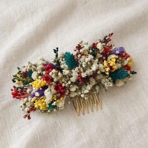 May include: A hair comb featuring a dense arrangement of dried flowers in vibrant colors including red, yellow, teal, and purple, interspersed with white baby's breath. The decorative floral cluster is attached to a gold-toned metal comb base.