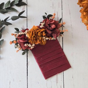 May include: A burgundy fabric rectangle with a floral arrangement of dried orange and red flowers on top. The flowers are arranged in a cluster and have small white flowers interspersed throughout.