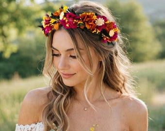 May include: A floral crown featuring a vibrant arrangement of deep magenta, burnt orange, and bright yellow artificial flowers interspersed with small green leaves and cream-colored accents, worn as a circular headpiece across the forehead.