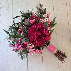 May include: A bouquet of dried flowers featuring deep magenta hydrangea blooms, light pink baby's breath, and dark green eucalyptus leaves. The stems are gathered together and wrapped in a bright pink fabric ribbon with a natural burlap trim.