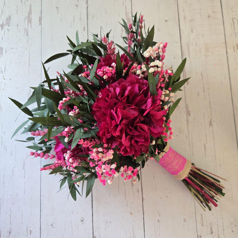 May include: A bouquet of dried flowers featuring deep magenta hydrangea blooms, light pink baby's breath, and dark green eucalyptus leaves. The stems are gathered together and wrapped in a bright pink fabric ribbon with a natural burlap trim.