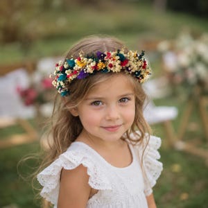 May include: A floral crown featuring a vibrant arrangement of dried flowers in shades of red, yellow, purple, and teal, accented with cream-colored baby's breath. The circular headpiece is constructed from small, delicate blossoms and foliage.