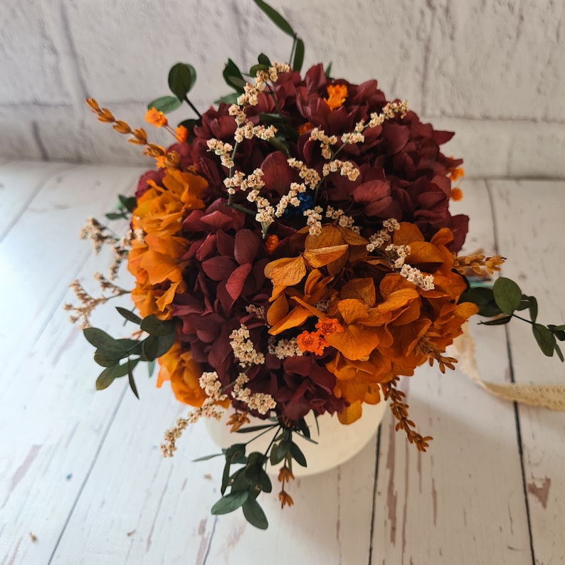 May include: A bouquet of dried hydrangeas in shades of burgundy and orange with small white flowers and green leaves.