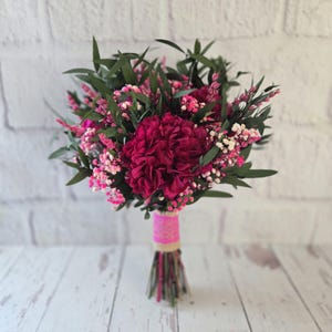 May include: A floral bouquet featuring deep magenta hydrangea blooms, small pink and white baby's breath, and dark green foliage. The stems are gathered together and wrapped in a band of tan burlap fabric with a bright pink decorative ribbon overlay.
