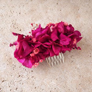 May include: A hair comb featuring a cluster of deep magenta preserved hydrangea petals and small light pink dried flowers. The decorative floral arrangement is attached to a silver-toned metal comb with multiple thin, curved teeth for securing.
