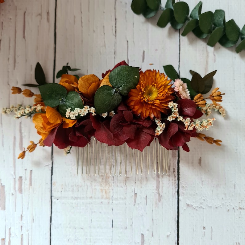 May include: A burgundy and orange floral hair comb with a silver comb. The comb is decorated with dried flowers, including burgundy hydrangeas, orange and yellow daisies, and small white flowers.