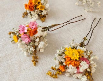 Könnte beinhalten: Drei florale Haarnadeln mit bunten Trockenblumen. Jede Nadel hat einen Cluster aus weißem Schleierkraut, akzentuiert mit gelben, orangefarbenen und rosa Blüten. Die Nadeln sind aus dunklem Metall und mit Draht umwickelt.