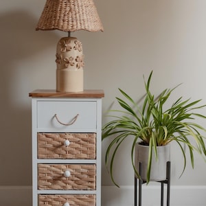 May include: A white bedside table with three woven basket drawers and a wooden top. A beige lamp with a woven shade and floral base sits on top. A potted plant with long green leaves is to the side.