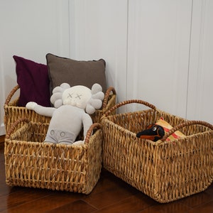 May include: Three woven wicker storage baskets of varying sizes. The baskets contain plush toys and pillows in shades of gray, purple, and brown. The baskets have brown handles and are placed against a white wall.