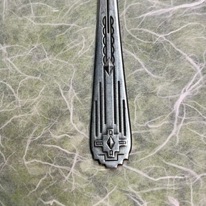 Puede incluir: Una pieza de cubiertos de plata con un intrincado diseño geométrico. El diseño presenta una serie de líneas verticales, una forma de diamante y un patrón repetitivo de círculos.