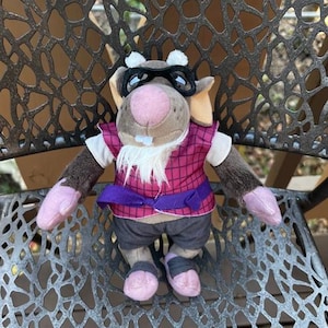 May include: A plush toy rat wearing glasses, a pink and purple vest, and gray pants. The rat has a pink nose, ears, and feet. It has a white beard and is sitting on a decorative metal chair.