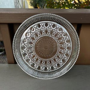 May include: Clear glass plate set with intricate embossed patterns. The set includes a large plate with a ribbed edge and a smaller plate with a floral-like design in the center. The plates are round and appear to be vintage.