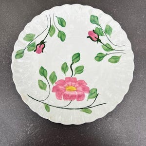 May include: A white ceramic plate with a scalloped edge features a hand-painted floral design. The design includes a large pink flower with a yellow center and smaller pink flowers with green leaves.