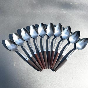 May include: A collection of nine silver spoons arranged in a fan shape. The spoons have a shiny, reflective surface with ornate detailing on the handles. The handles transition to a dark brown color.